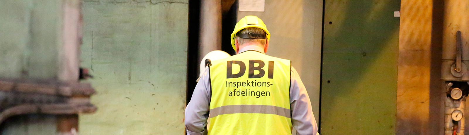 DBI-medarbejder med gul sikkerhedshjelm og refleksvest mærket ”DBI Inspektionsafdelingen” ved store maskiner.