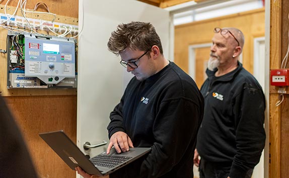 To AMU-kursister undersøger et åbent brandalarmeringsanlæg, mens den ene arbejder på en bærbar computer.