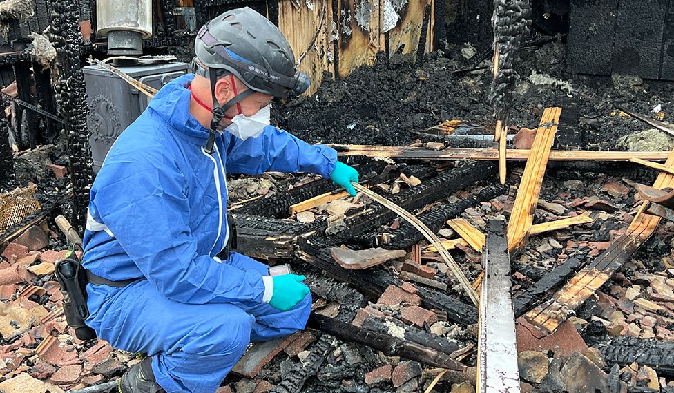 Brandundersøger undersøger forkullede rester og terrassebrædder ved nedbrændt sommerhus.